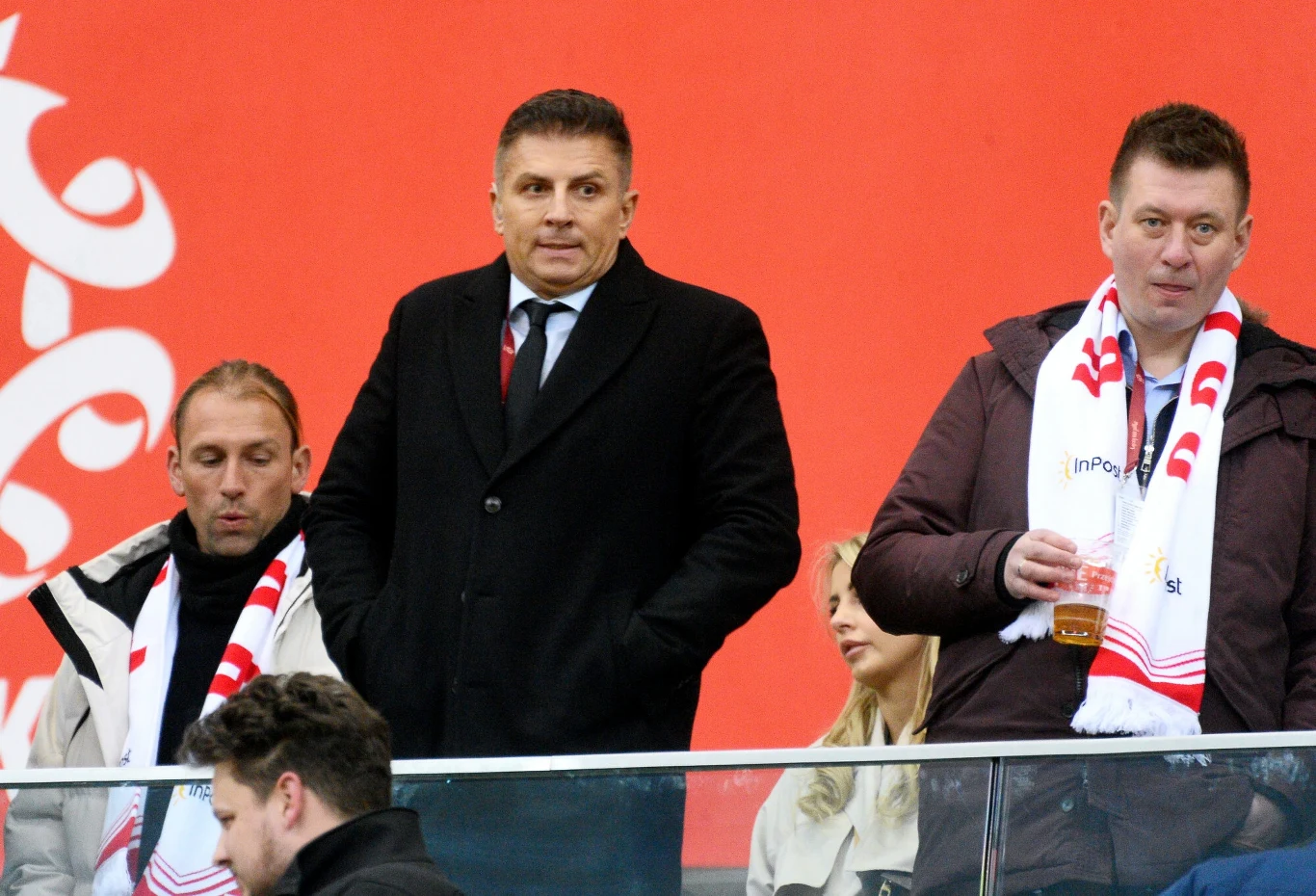 Kilku mężczyzn ubranych w eleganckie płaszcze i szaliki z biało-czerwonym motywem stoi w loży stadionowej, jeden z nich trzyma plastikowy kubek z napojem, a w tle znajduje się czerwona ściana z fragmentami białego napisu.