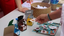 Osoba trzymająca kartonik z obrazkiem nad stołem, na którym znajdują się różne karty do gry, kolorowe pudełka na karty oraz koszyk z kartkami papieru, wokół widoczne ręce innych uczestników.