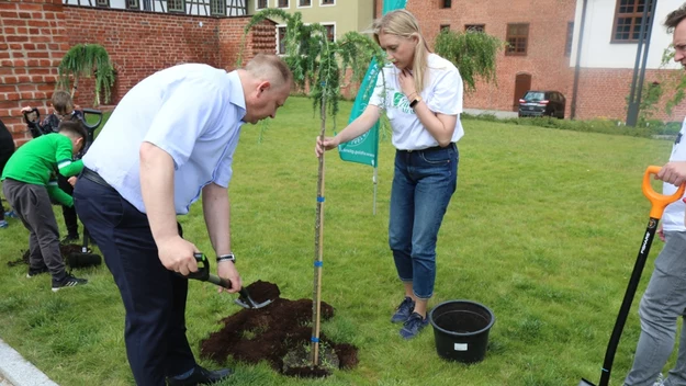 Mężczyzna sadzi młode drzewko na trawniku, kobieta w białej koszulce przytrzymuje drzewko, w tle dzieci i inne osoby uczestniczące w akcji sadzenia oraz budynki z cegły.