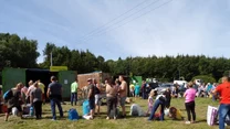 Grupa osób ustawia się z workami i torbami w kolejce przy kontenerach na odpady, otoczenie stanowi teren zielony z drzewami i trawą, w tle widoczne są samochody i oznaczenia do segregacji śmieci.