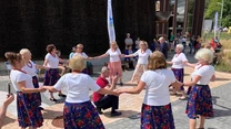 Grupa starszych kobiet w białych bluzkach i kolorowych spódnicach tworzy krąg i trzyma się za ręce, tańcząc na świeżym powietrzu, wokół widzowie i przechodnie obserwują ich występ.