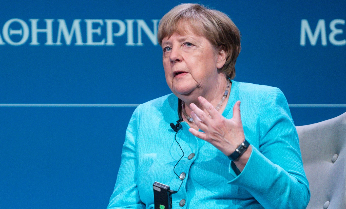 Angela Merkel nie wycofuje się ze swojego poparcia dla migracji Kobieta w średnim wieku w niebieskim żakiecie siedzi na scenie z mikrofonem przypiętym do ubrania, wykonując gest dłonią podczas rozmowy lub przemówienia, w tle niebieskie tło z białymi napisami.