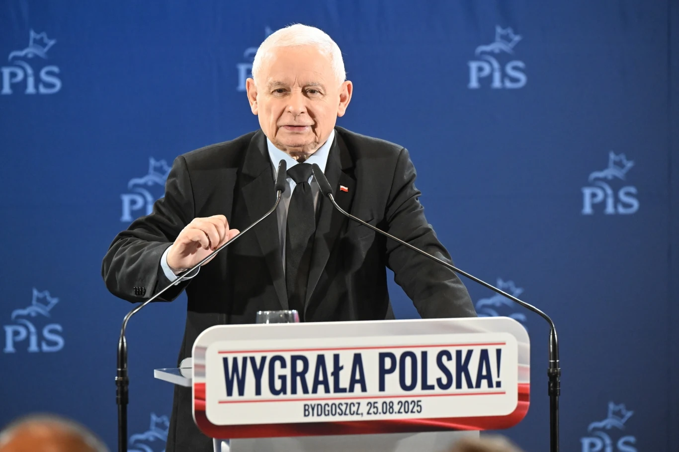 Prezes PiS Jarosław Kaczyński na spotkaniu w Bydgoszczy Polityk stojący za mównicą z napisem 'Wygrała Polska!' oraz datą i miejscowością, na tle granatowej ścianki z logotypami partii PiS, ujęcie z eventu politycznego.