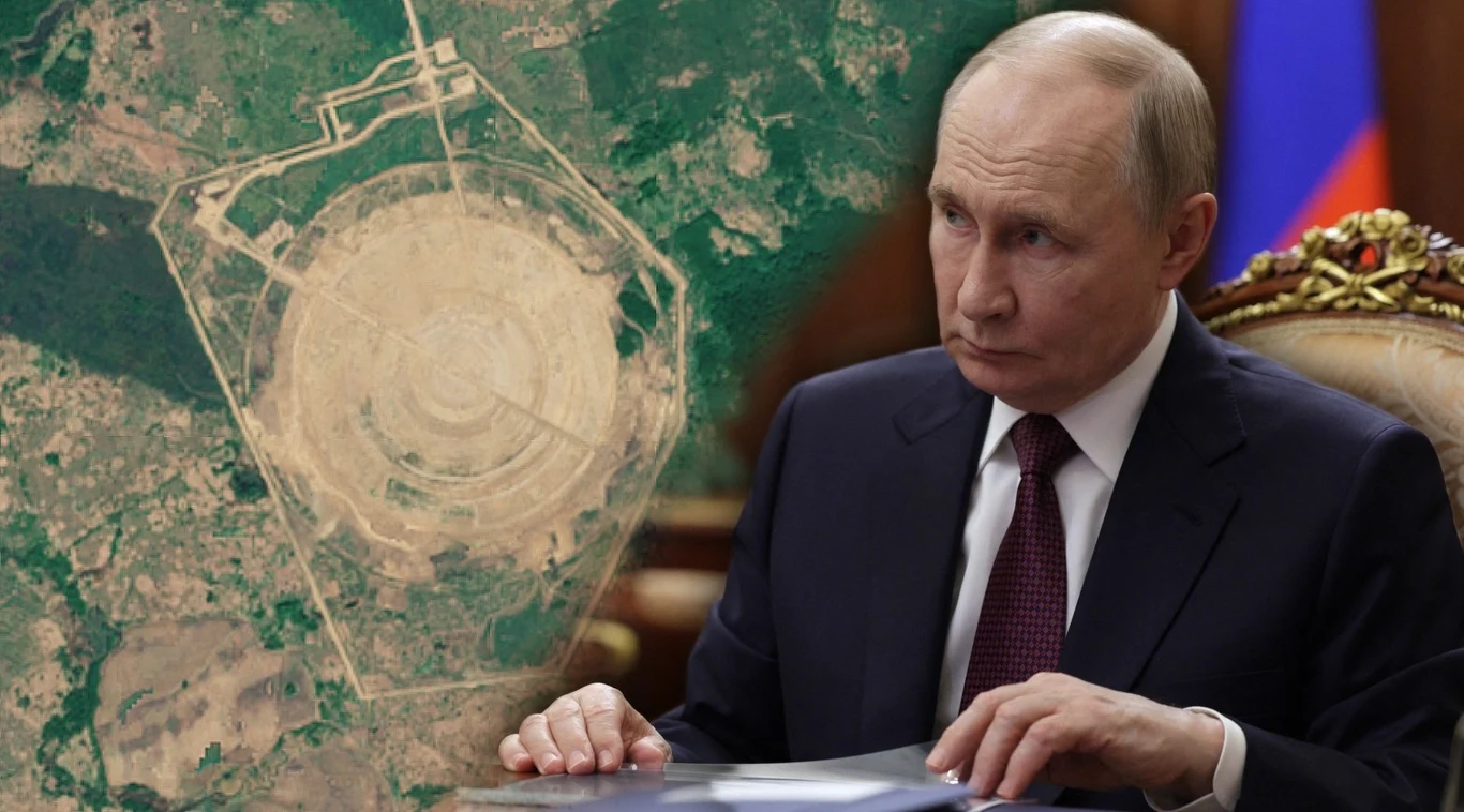 Zdjęcie satelitarne rosyjskiego kompleksu wykonane przez satelity systemu Copernicus Prezydent Rosji Władimir Putin siedzący przy stole, w tle widoczny satelitarny obraz dużej instalacji o zaawansowanej strukturze rozmieszczonej na rozległym terenie; flaga Rosji w tle po prawej stronie.
