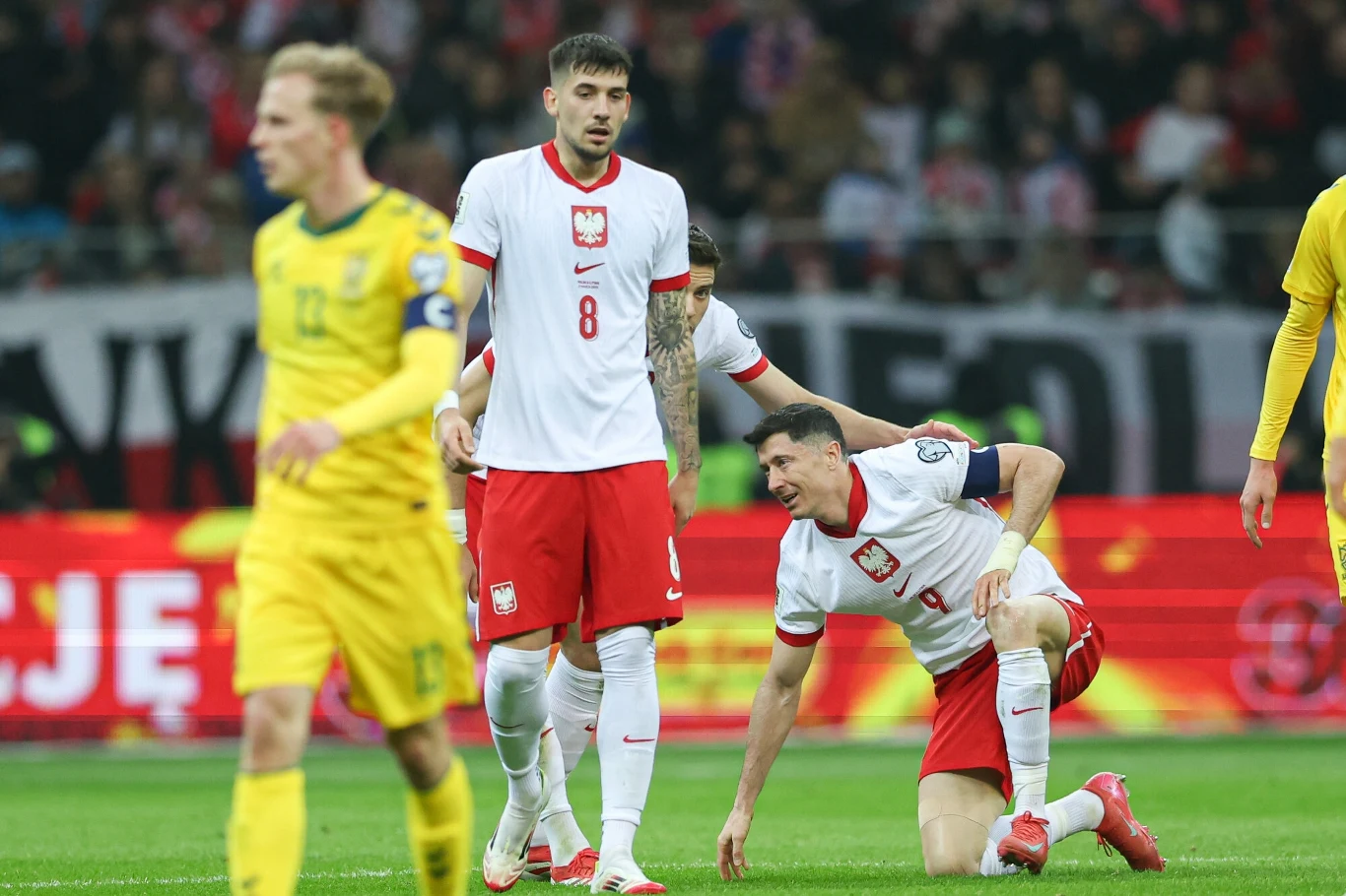 Dwaj polscy piłkarze w białych koszulkach i czerwonych spodenkach podczas meczu, jeden z nich wspiera drugiego, który klęczy na boisku, obok zawodnik rywali w żółtym stroju, a w tle trybuny wypełnione kibicami.