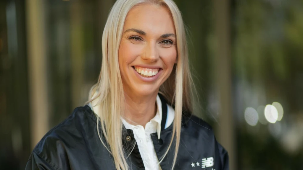 Iga Baumgart-Witan Uśmiechnięta kobieta o długich blond włosach, ubrana w czarno-białą sportową kurtkę, stoi na tle rozmytego, zielonego otoczenia.