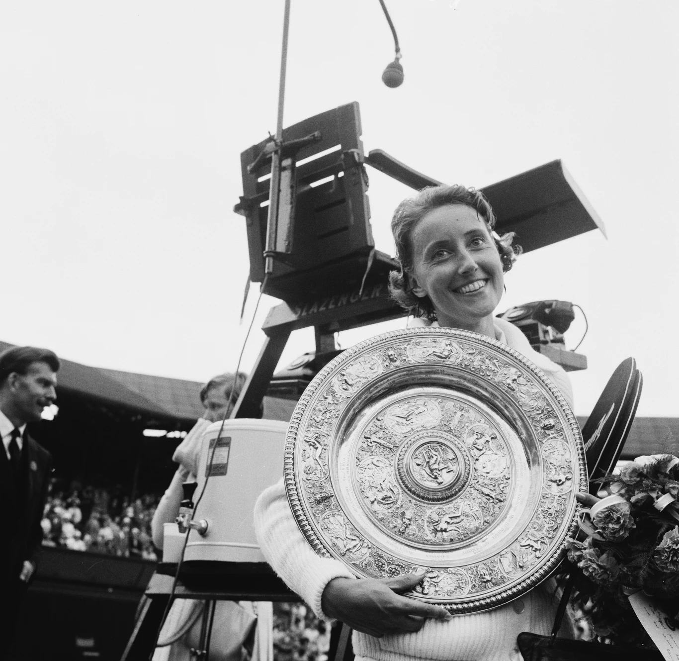 Uśmiechnięta tenisistka trzyma w rękach trofeum w kształcie srebrnej tacy, obok bukiet kwiatów, w tle widoczna kamera telewizyjna oraz częściowo zasłonięte osoby.