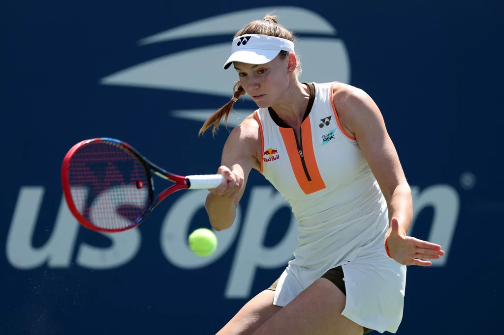 Tenisistka w białej sukience sportowej odbijająca piłkę podczas meczu tenisowego, w tle widoczne logo turnieju US Open.