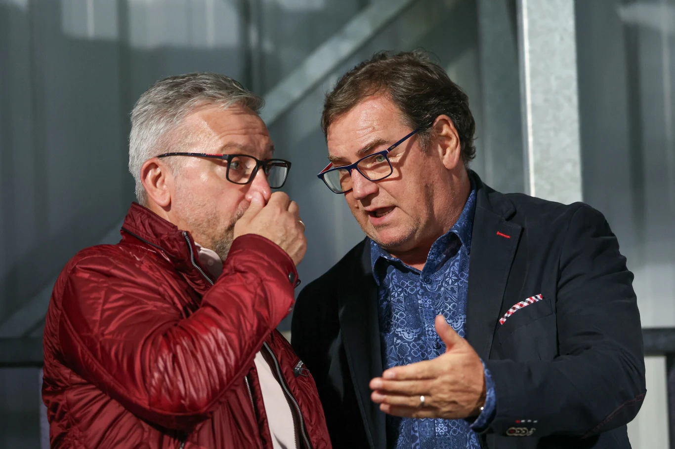 Jan Urban i Jerzy Brzęczek Dwóch mężczyzn w średnim wieku prowadzi poważną rozmowę, jeden z nich ma okulary i granatową marynarkę, drugi czerwono-brązową kurtkę i przykłada rękę do ust, oboje stoją w jasnym wnętrzu.