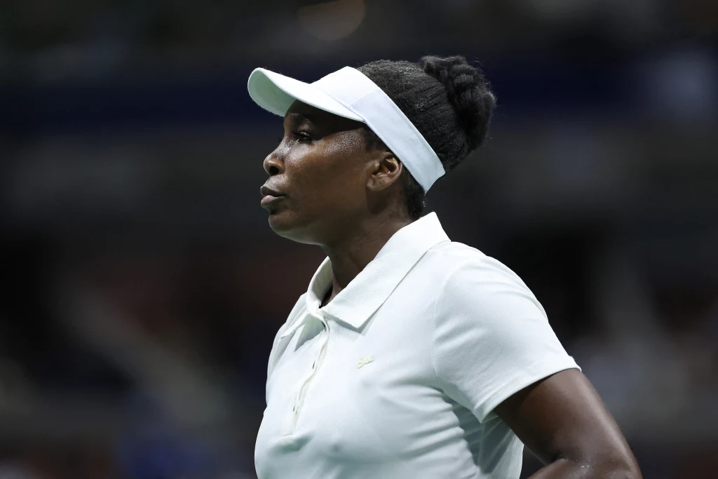 Kobieta w sportowym stroju i białej czapce z daszkiem na tle rozmytego stadionu, skupiona i poważna, podczas wydarzenia sportowego.