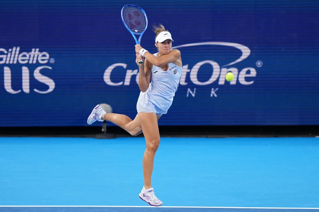 Tenisistka uderzająca piłkę na błękitnym korcie, ubrana w jasny strój sportowy i czapeczkę, w tle widoczne reklamy sponsorów.