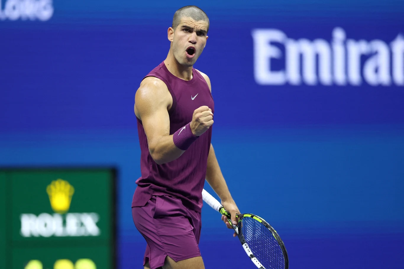 Tenisista w fioletowym stroju wykonuje gest triumfu z zaciśniętą pięścią, stojąc na korcie z rakietą w lewej ręce, w tle widoczne logo sponsora oraz tablica wyników.