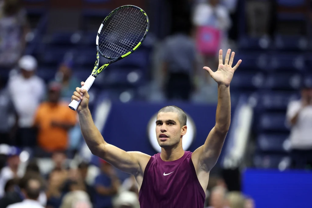 Młody tenisista w sportowej koszulce z podniesionymi obiema rękami, w jednej trzyma rakietę tenisową, wokół widoczni kibice i trybuny stadionu.