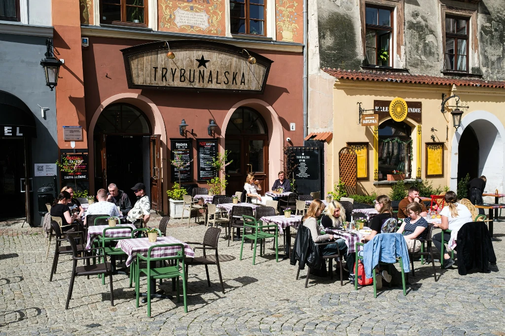 Ludzie siedzą przy stolikach na zewnątrz kawiarni na miejskim rynku, rozmawiają i jedzą posiłki, otoczeni kolorowymi elewacjami kamienic, atmosfera jest spokojna i przyjazna.