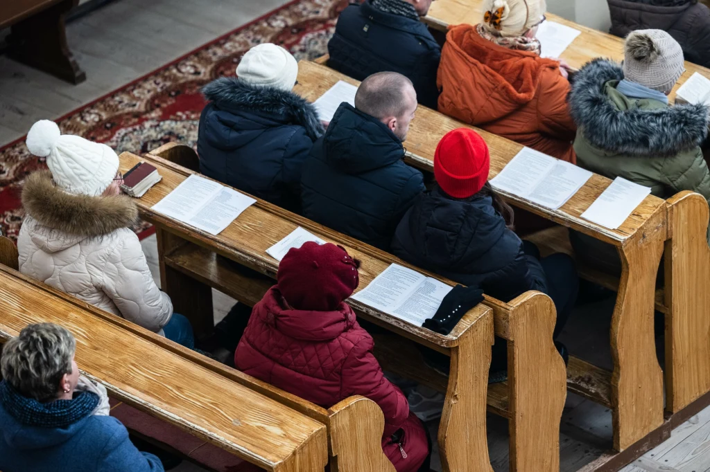 Grupa osób siedząca w ławkach w kościele, każdy z nich ma przed sobą otwartą książkę, większość ubrana w zimowe kurtki i czapki, widok z góry.