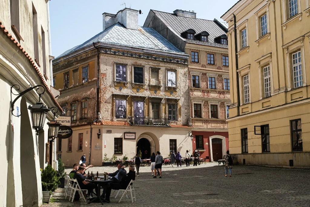 Zabytkowa kamienica z odnowioną fasadą, na której widoczne są czarno-białe fotografie, otoczona innymi budynkami, przy których ustawione są stoliki z siedzącymi ludźmi, w tle chodzący przechodnie na brukowanej ulicy.