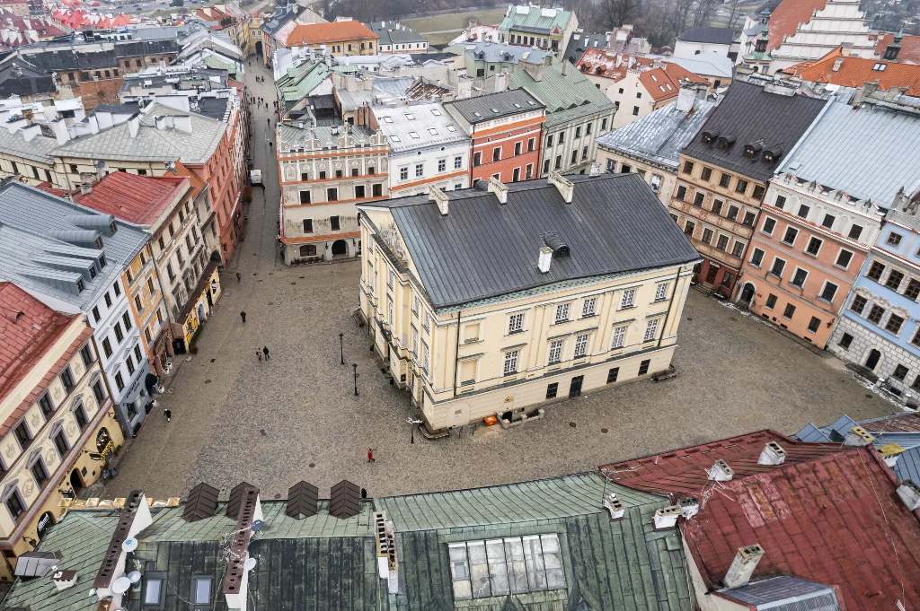 Rynek otoczony zabytkowymi kamienicami o różnokolorowych dachach, pośrodku placu centralnie ustawiony większy budynek, brukowana powierzchnia placu i niewielu przechodniów widocznych z góry.
