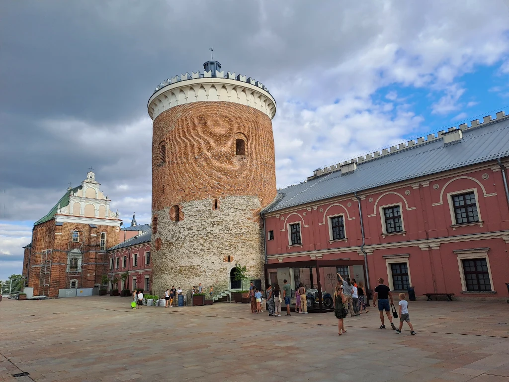 W centrum kadru znajduje się okrągła, ceglana wieża otoczona historycznymi budynkami o różowej elewacji po prawej oraz bardziej klasycznym, jasnym budynku po lewej stronie. Na placu przed wieżą zgromadzone są grupy turystów zwiedzających zabytek, część...
