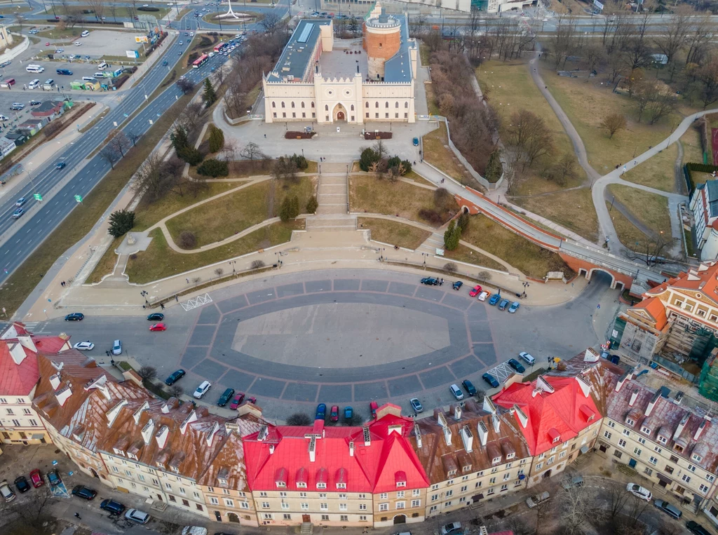 Panorama lubelskiego zamku z góry, przed którym rozpościera się szeroki plac i półkoliste kamienice z czerwonymi dachami, otoczone przez ulice oraz parkowe alejki.