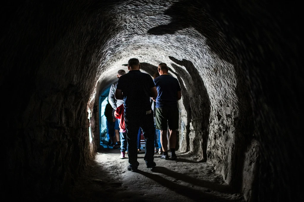 Grupa osób idąca wąskim, oświetlonym korytarzem skalnym z wyjściem oświetlonym w tle, ściany tunelu mają chropowatą fakturę a światło tworzy wyraziste cienie na podłożu.