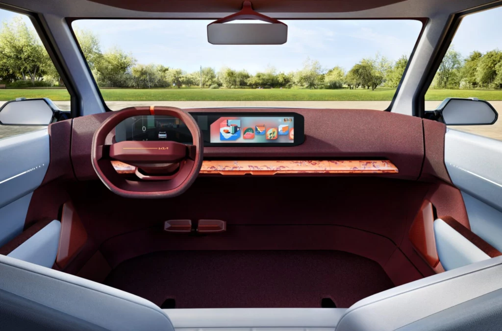 Produkcja EV2 ruszy w lutym 2026 r. w zakładzie w Żylinie, gdzie już teraz powstaje elektryczny model EV4 oraz spalinowe Sportage i XCeed. Minimalistyczne, nowoczesne wnętrze samochodu z elektronicznym panelem deski rozdzielczej i dużym panoramicznym ekranem, widok z perspektywy tylnego siedzenia na kierownicę oraz przestrzeń przednią, za oknem widać zielony krajobraz.