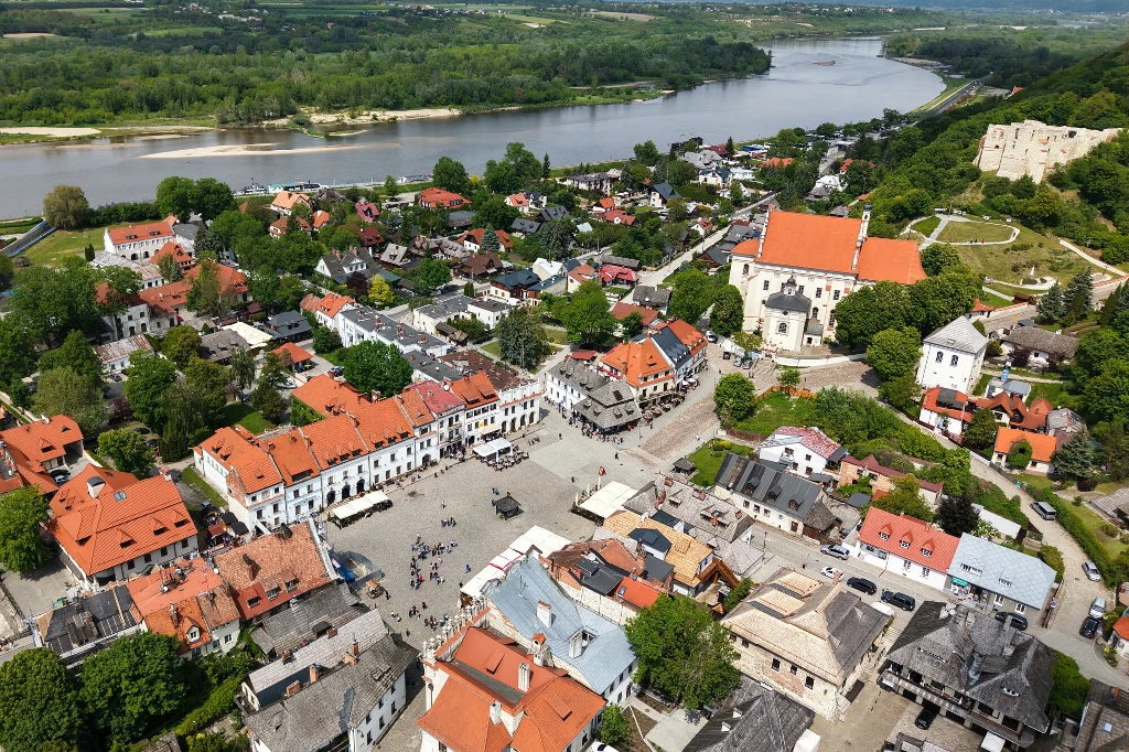 Panorama małego miasta z charakterystyczną zabudową o czerwonych dachach, widoczny rynek otoczony kamienicami, zabytkowy kościół i ruiny zamku na wzgórzu, w tle rzeka oraz zielone tereny.