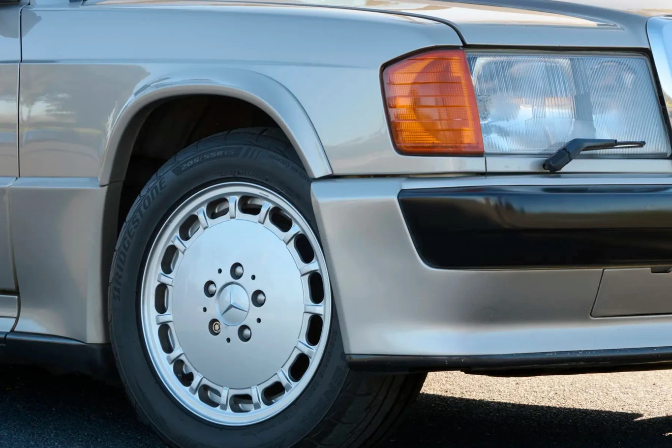 Mercedes Ayrtona Senny wystawiony na sprzedaż | Fot. RM Sotheby's Przedni lewy reflektor, zderzak i koło klasycznego srebrnego samochodu marki Mercedes-Benz, widoczne detale felgi oraz fragment karoserii.