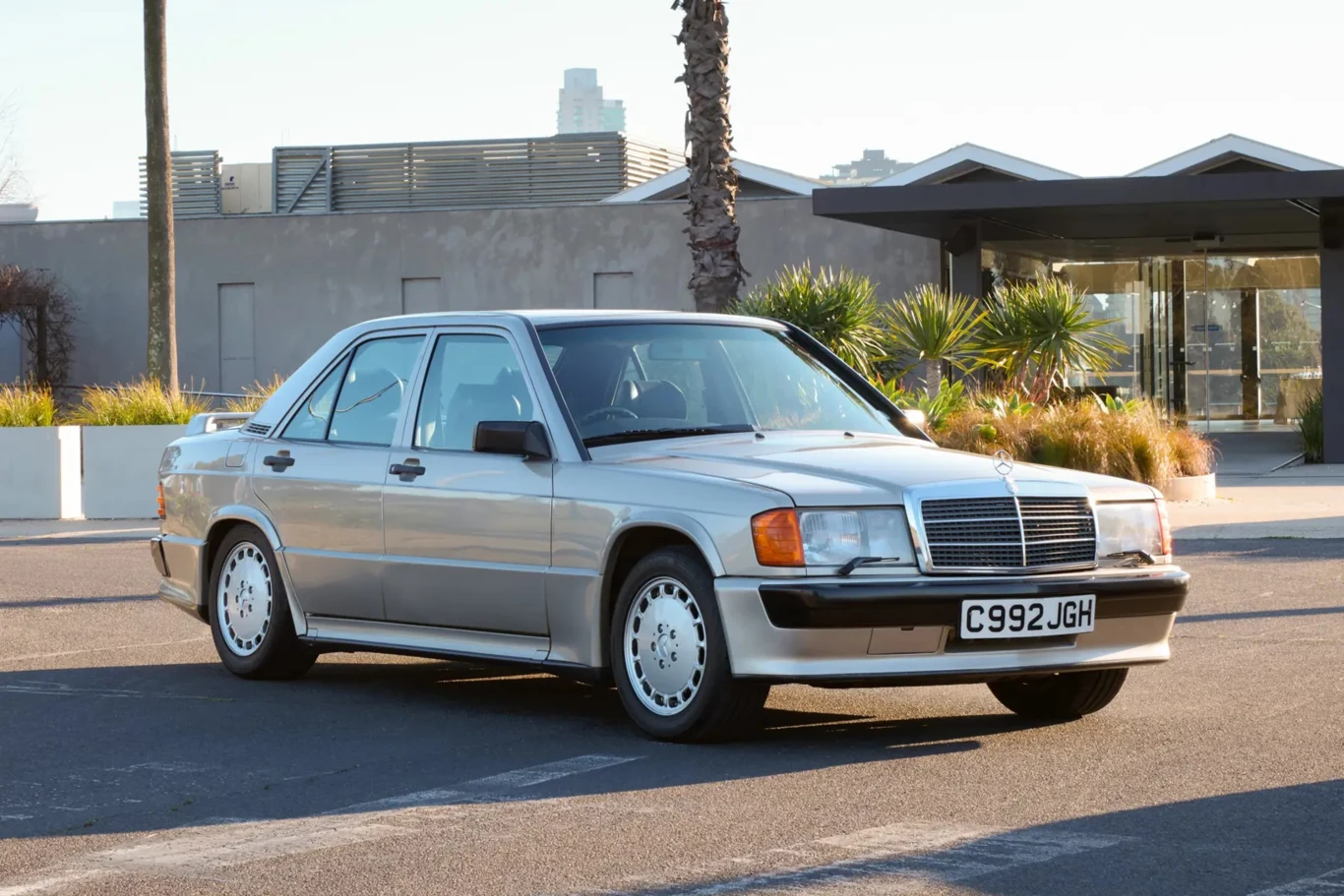 Za kierownicą podobnego Mercedesa, Senna olśnił cały świat. | Fot. RM Sotheby's Srebrny samochód osobowy marki Mercedes-Benz z lat 80. zaparkowany na ulicy, w tle nowoczesna architektura, palma i roślinność.