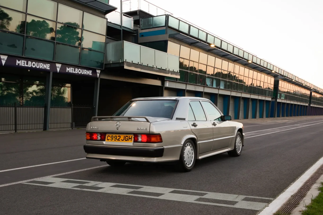 Senna zamówił tego Mercedesa prosto z fabryki w 1985 roku. | Fot. RM Sotheby's klasyczny srebrny sedan marki Mercedes-Benz jadący po pustej nitce toru wyścigowego obok nowoczesnych trybun z przeszklonymi balkonami oraz widoczną linią mety na pierwszym planie