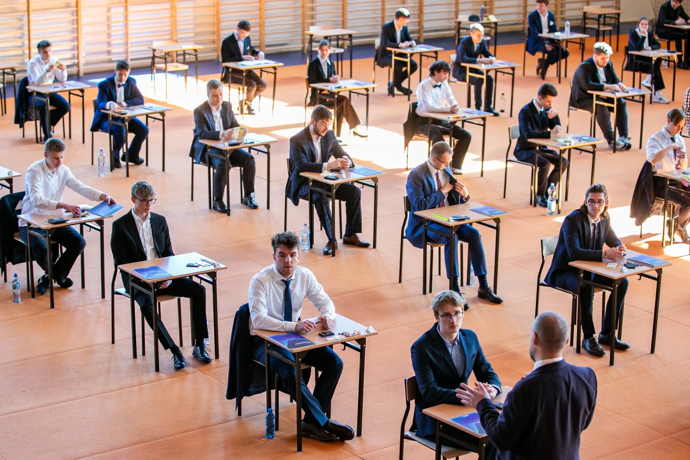 Kiedy będą matury 2026? Prezentujemy harmonogram CKE Uczniowie siedzący przy pojedynczych ławkach w dużej sali gimnastycznej, ubrani elegancko, rozdzieleni od siebie na bezpieczną odległość, przed nimi arkusze egzaminacyjne, a nauczyciel na pierwszym planie tłumaczy zasady lub motywuje przed rozpoczęciem...