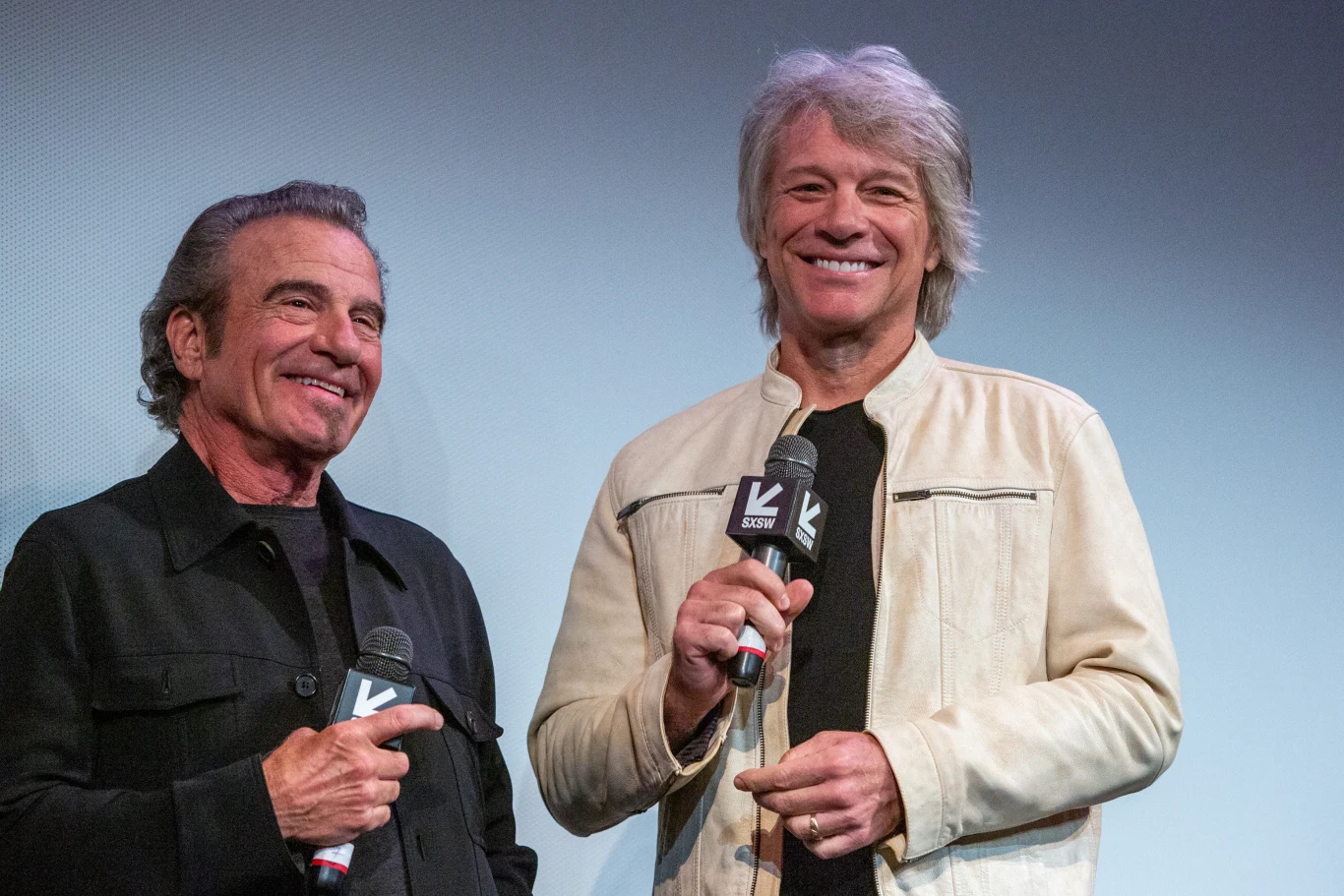 Tico Torres i Jon Bon Jovi występują razem pod szyldem Bon Jovi od 1983 r. Dwóch uśmiechniętych mężczyzn trzymających mikrofony na tle neutralnego, lekko niebieskiego tła, jeden ubrany w czarną kurtkę, drugi w jasną kurtkę, obaj stoją blisko siebie i patrzą w stronę publiczności.