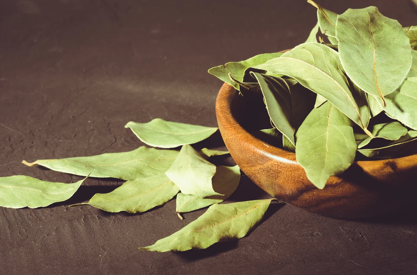 Drewniana miska wypełniona liśćmi laurowymi, kilka liści rozsypanych na ciemnym tle obok miski.
