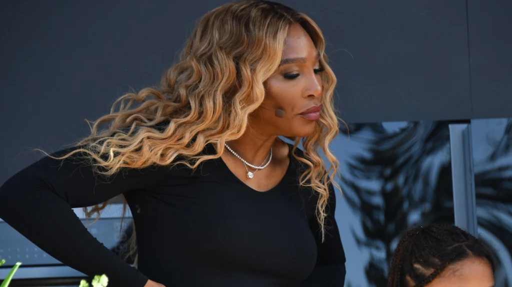 Kobieta o długich falowanych blond włosach ubrana na czarno, z delikatnym makijażem i naszyjnikiem, patrzy w bok z poważnym wyrazem twarzy.