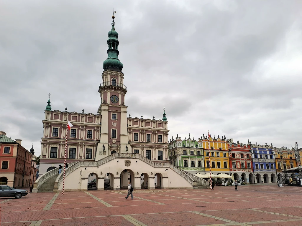 Ratusz w Zamościu z charakterystyczną wieżą i ozdobnymi schodami na centralnym placu rynku, otoczony kolorowymi kamienicami pod pochmurnym niebem.