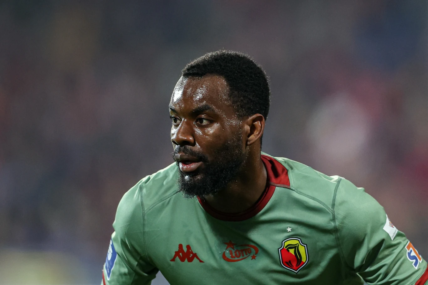 Piłkarz w zielonej koszulce z herbem klubu oraz logotypami sponsorów podczas gry, z wyrazem skupienia na twarzy, na rozmytym tle stadionu.