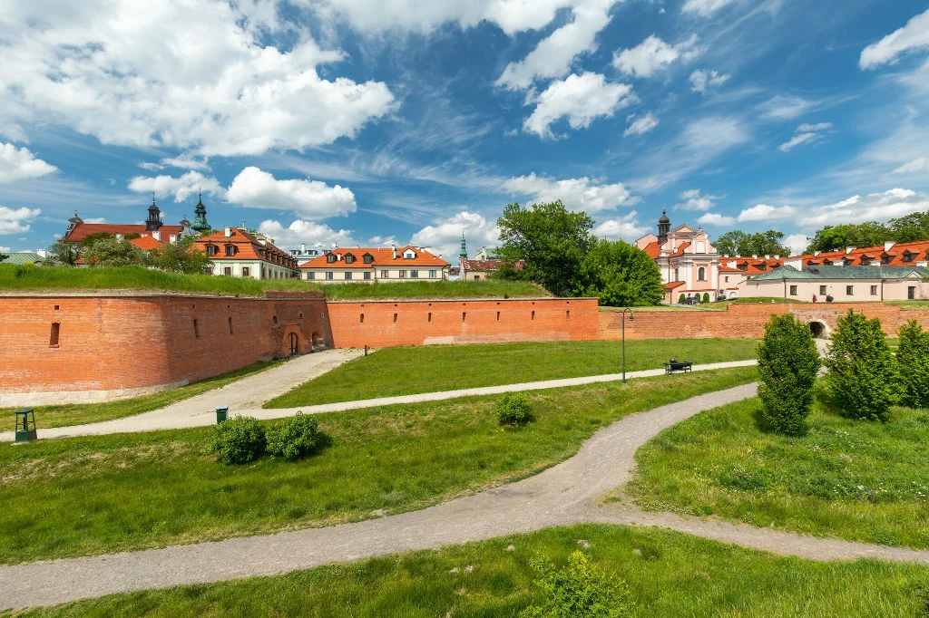Ceglane mury twierdzy otoczone zielonym trawnikiem, w tle zabudowania z czerwonymi dachami i wieże kościoła pod błękitnym niebem z białymi chmurami.