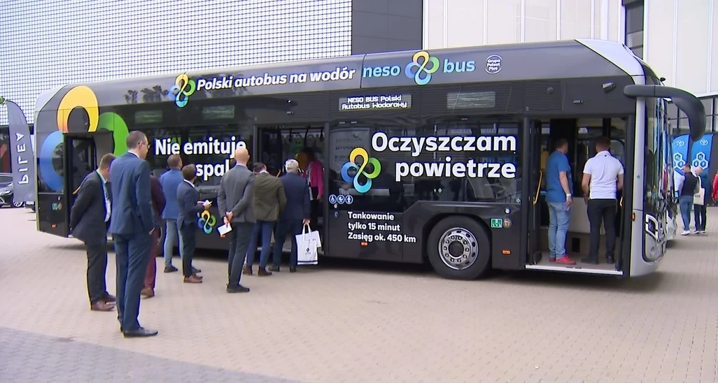 NesoBus Fot. Polsat News Hydrogenowy autobus miejski neso bus otoczony przez grupę osób, którzy oglądają i wchodzą do pojazdu. Na pojeździe widoczne napisy informujące o jego ekologicznych zaletach, takich jak brak emisji spalin i oczyszczanie powietrza.