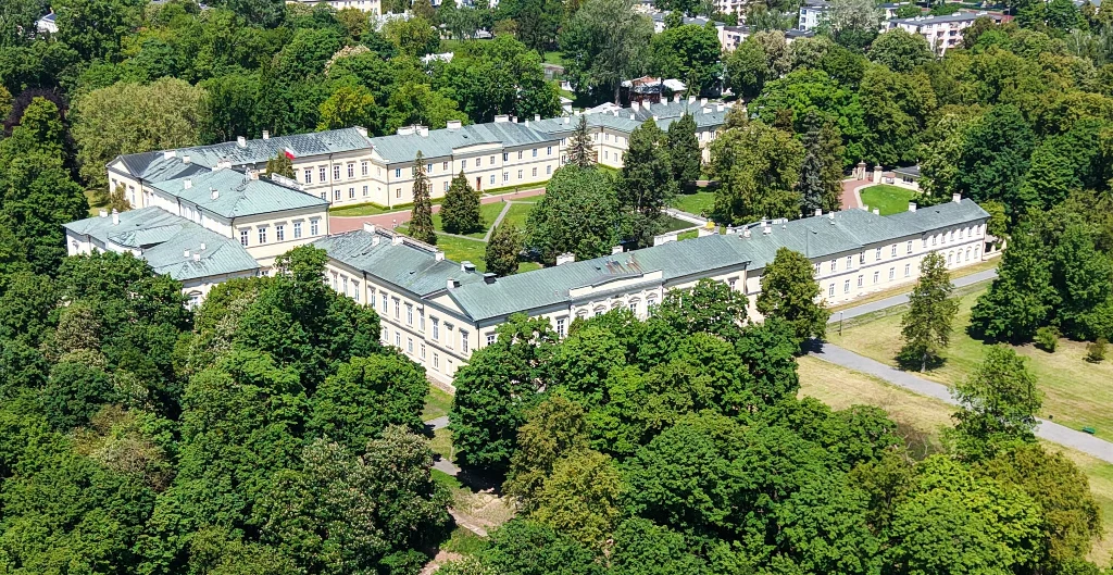 Zabytkowy kompleks pałacowy z jasnymi budynkami i zielonym dachem położony w centrum rozległego, zadrzewionego parku, otoczony wysokimi drzewami i alejkami spacerowymi.