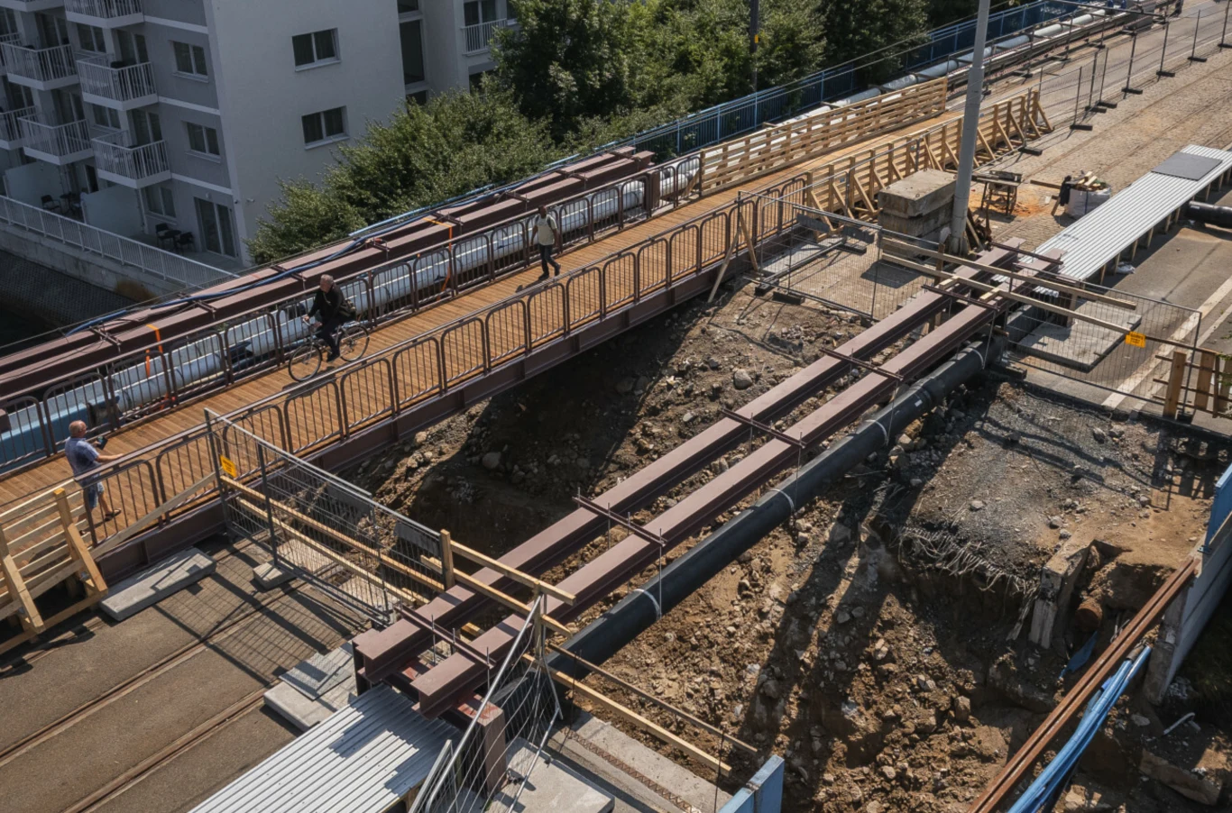 Most Siennicki w remoncie, otwarty dla pieszych i rowerzystów. Konstrukcja mostu nad głębokim wykopem ziemnym, po której przechodzą piesi i rowerzysta, otoczona zabezpieczeniami i barierkami, obok znajduje się przystanek tramwajowy i zabudowania mieszkalne.