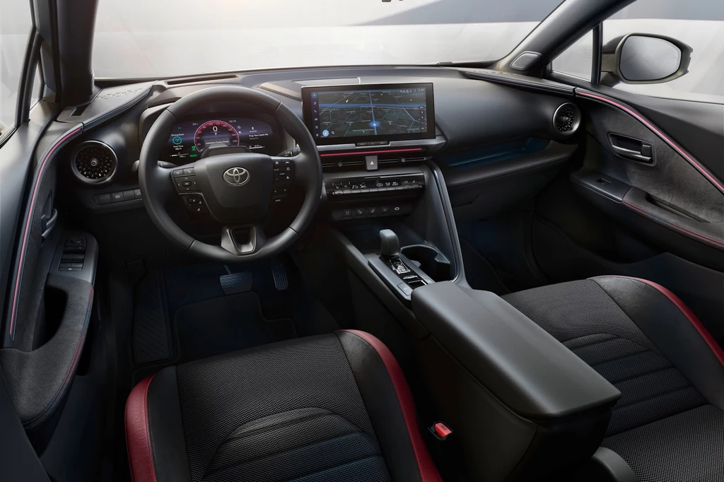 Nowoczesne wnętrze samochodu marki Toyota z cyfrowymi wskaźnikami, dużym wyświetlaczem na konsoli środkowej oraz sportowymi fotelami obitymi czarną tapicerką z czerwonymi akcentami.
