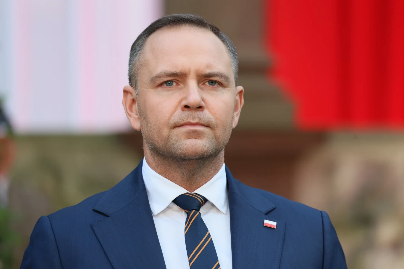 Mężczyzna w garniturze i krawacie z przypiętą flagą Polski na tle rozmytych barw czerwono-białych, wyrażający powagę i skupienie.