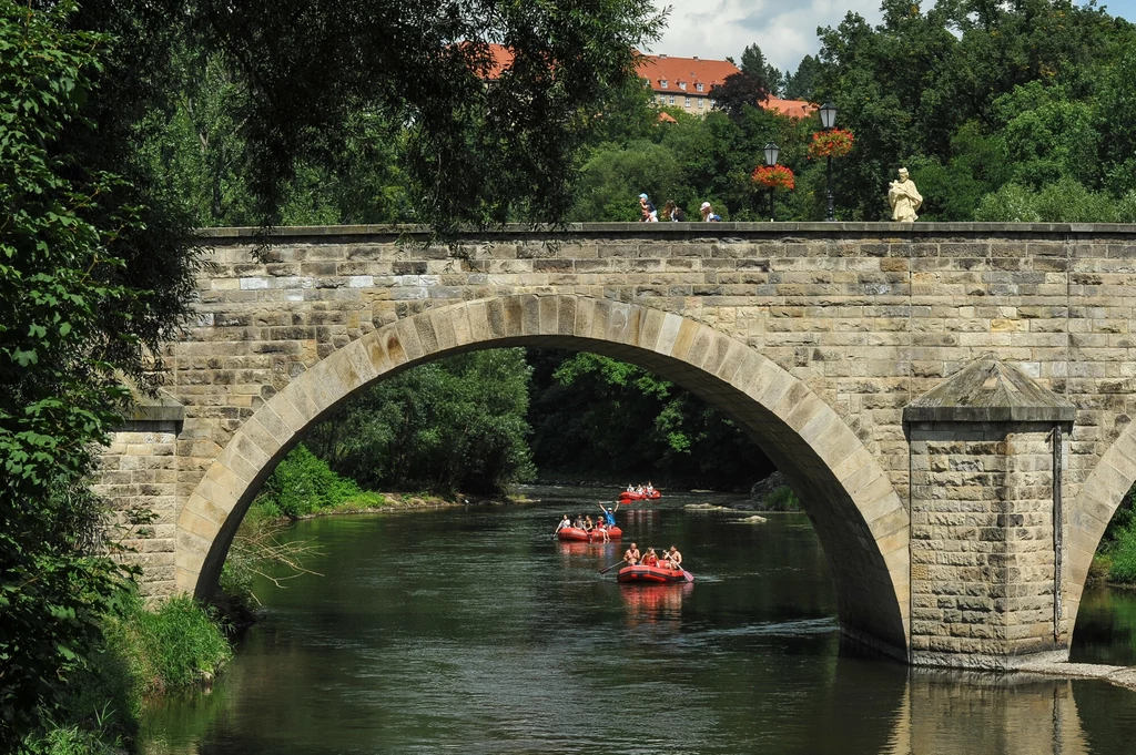 Spływ pontonowy to świetna przygoda dla całej rodziny Kamienny most łukowy nad rzeką, pod którym płyną osoby na pontonach, w tle drzewa i fragmenty czerwonych dachów zabudowań.