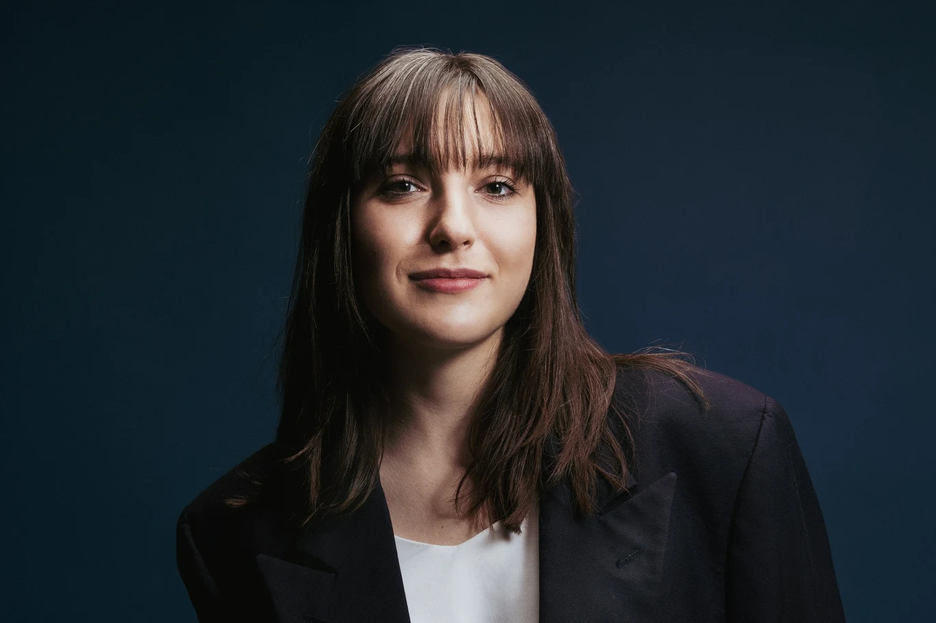 Julia Łozowska Młoda kobieta z prostymi, ciemnymi włosami i grzywką ułożoną na czoło, ubrana w ciemną marynarkę i jasną bluzkę, patrzy prosto w obiektyw na jednolitym, ciemnoniebieskim tle.