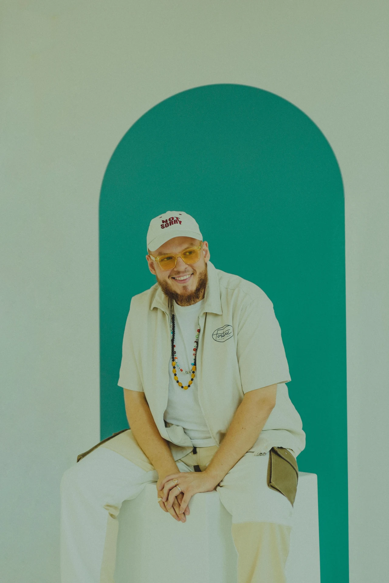 Wojciech Lechończak „Cieniu” Uśmiechnięty mężczyzna z brodą w jasnych ubraniach i czapce siedzi na białym postumencie, w tle widoczny turkusowy łuk.