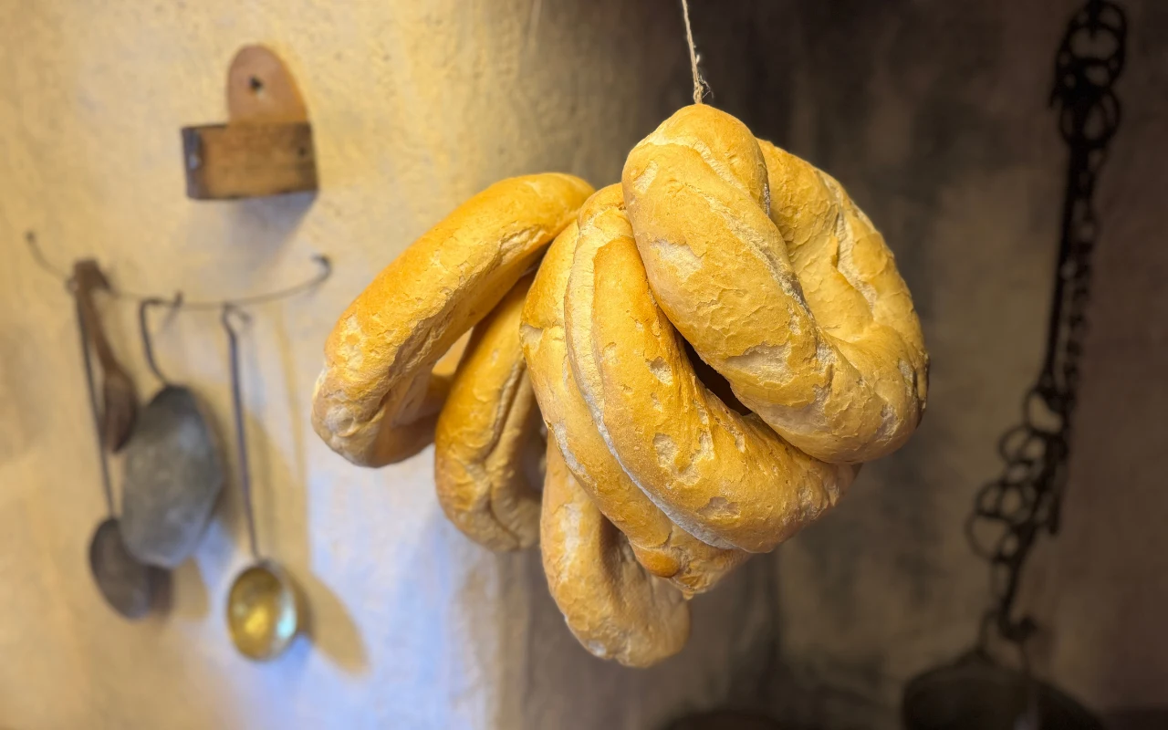 Kilka obwarzanków zawieszonych na sznurku w rustykalnej kuchni, w tle kuchenne narzędzia wiszące na ścianie.