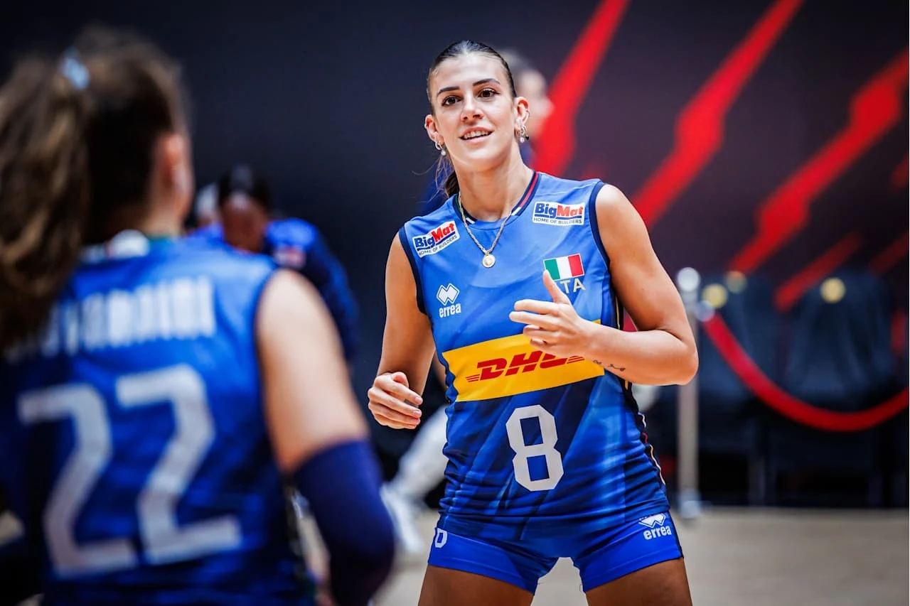 Siatkarka w stroju reprezentacji Włoch z numerem 8 na koszulce stoi na hali sportowej podczas meczu, obok druga zawodniczka z numerem 22 z widocznym tylko fragmentem twarzy. Tło stanowi ciemna ściana z czerwonymi elementami graficznymi.