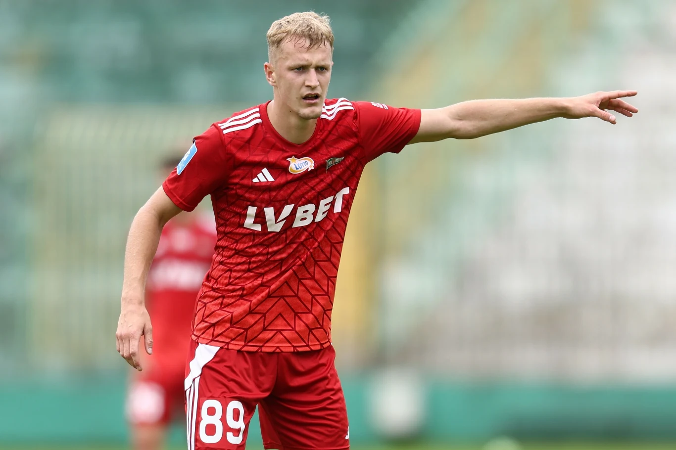 Piłkarz ubrany w czerwony strój sportowy z numerem 89 i logo sponsora stoi na boisku i gestykuluje, wskazując ręką kierunek gry podczas meczu.