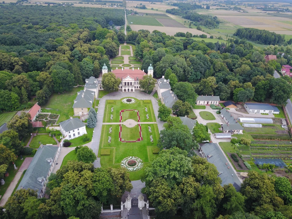 Rezydencja pałacowa otoczona rozległymi, symetrycznie zaprojektowanymi ogrodami francuskimi, liczne alejki, żywopłoty i rabaty kwiatowe, budynki gospodarcze oraz gęsty las w tle, w oddali rozległe pola uprawne.