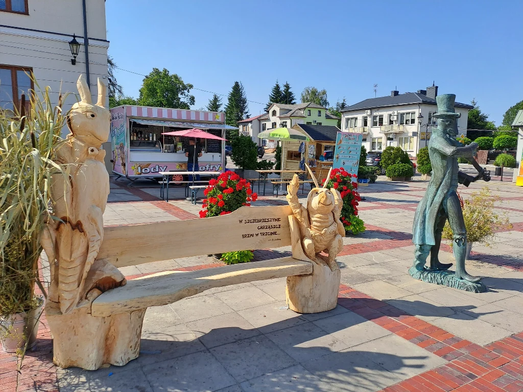 Drewniana ławka ozdobiona rzeźbami postaci z bajki, obok metalowa statua mężczyzny w cylindrze, w tle kwiaty, kolorowe stoiska i budynki miejskie.