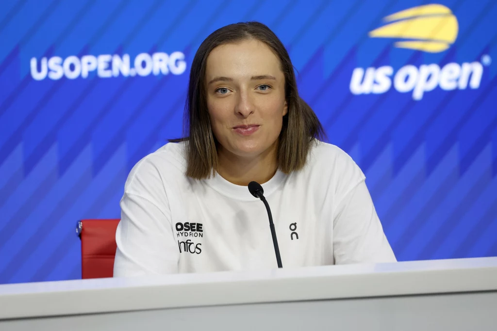 Iga Świątek na US Open Kobieta z krótkimi brązowymi włosami w białej koszulce siedzi przy stole konferencyjnym z mikrofonem, na tle niebieskiej planszy z logotypami turnieju US Open.